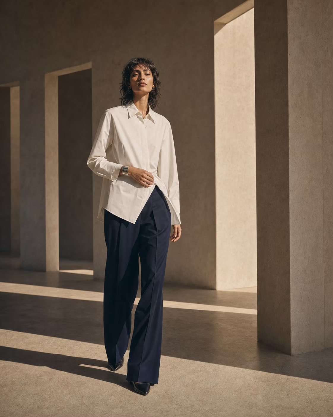 Model in an ivory shirt and navy trousers walking through a stone arcade.